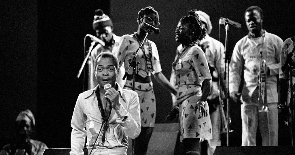1024x538_cmsv2_cf4578f6-42c4-525a-8b51-e6a979ed33fe-7132974.jpg Black and white photo of a man singing into a microphone on stage with women backup singers and a band with instruments behind him, reminiscent of Fela Kuti, the Nigerian King of Afrobeat.
