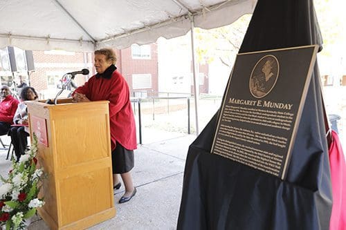 10861.jpg A woman stands at a podium speaking under a tent. In the foreground, a plaque dedicated to Margaret E. Munday, the first Black undergraduate pupil at WKU, is on display.