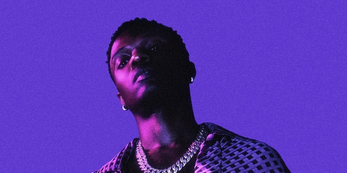 1667235035_wizkid.jpg A person with a serious expression, wearing a patterned shirt and chain necklace, stands against a solid purple background, reminiscent of the rich tones often seen in October.