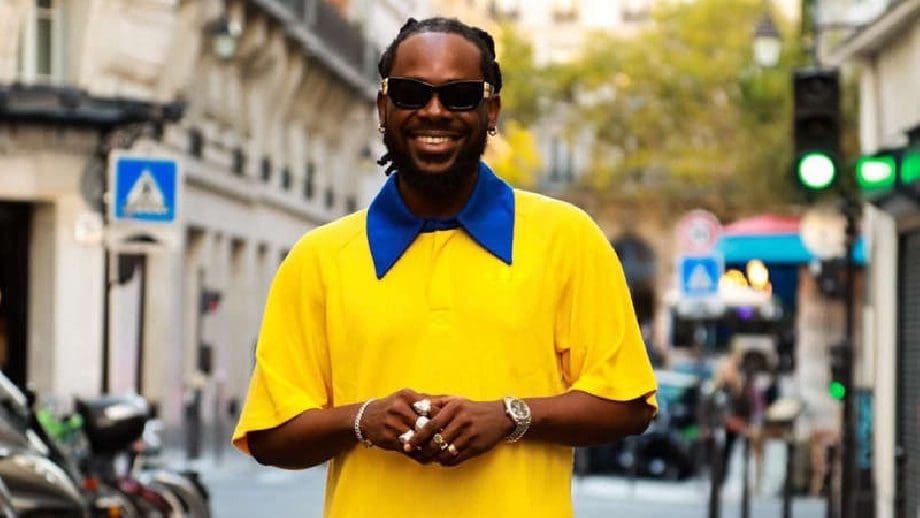 6357ee5b060f6.jpg A man wearing sunglasses and a yellow shirt with a blue collar stands smiling on a city street, perhaps reminiscing about the Finest Present Ever when he attended the Adekunle Gold Lagos concert.