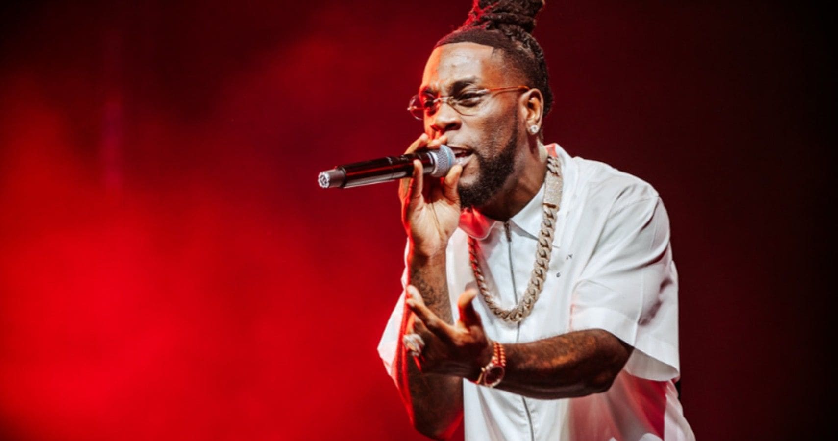 A-Picture-Of-Burna-Boy.jpg A man with dreadlocks and glasses performs passionately on stage, holding a microphone in his right hand. He is wearing a white shirt and multiple necklaces, set against a red-lit background. Among the richest Afrobeat artists, his energy captivates the crowd.