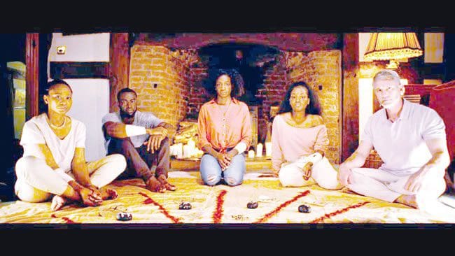 A-song-from-the-dark.jpg Five people sit on the floor around a patterned cloth with objects placed on it, against a backdrop of a brick wall with candles, creating an atmosphere that feels like a scene from "A Track from the Darkish.