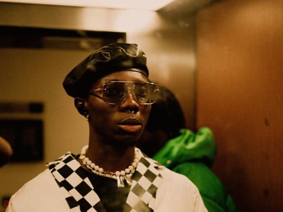 Blaqbonez-Video-Snippet-Young-Preacher-NotjustOK-1.jpeg Person wearing glasses and a black beret stands in an indoor setting in London. They are dressed in a checkered outfit with a white necklace. Another person in a green jacket is in the background.