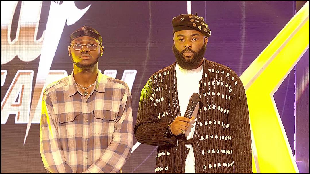 Contestant-Emzez-Host-of-the-Show-VJ-ADAMS.jpeg Two men stand side by side on a stage. One wears a plaid shirt and cap; the other, in a patterned cardigan and hat, holds a microphone. A partially visible sign is in the background as Afrobeats rhythms pulse through the air, introducing new sounds to an eager audience.