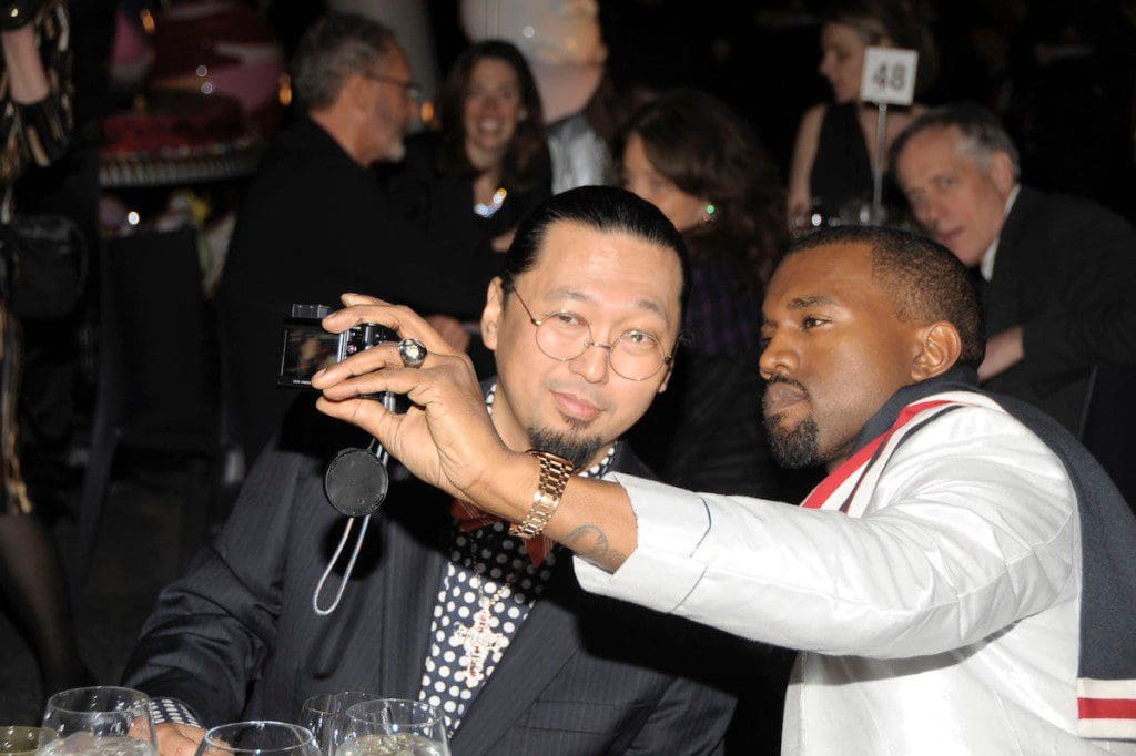 GettyImages-620277572.jpg Two men sitting at a table take a selfie at an event, one holding a camera. Amongst the crowd of artist collaborators, they capture the moment. Other people are visible in the background.