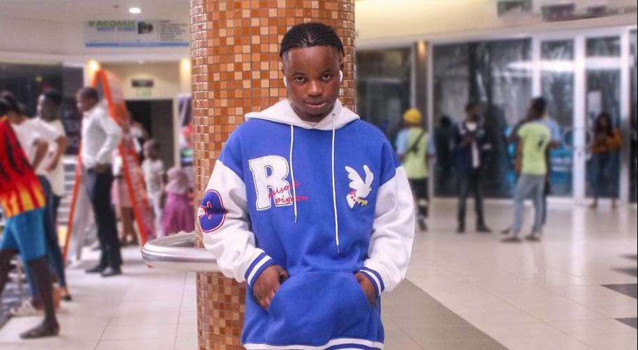 Kaptain-Kush-e1666458668728.jpeg A young man wearing a blue and white hoodie, reminiscent of a Style Of Fame look, stands with his hands in his pockets in a bustling indoor space with several other people in the background.