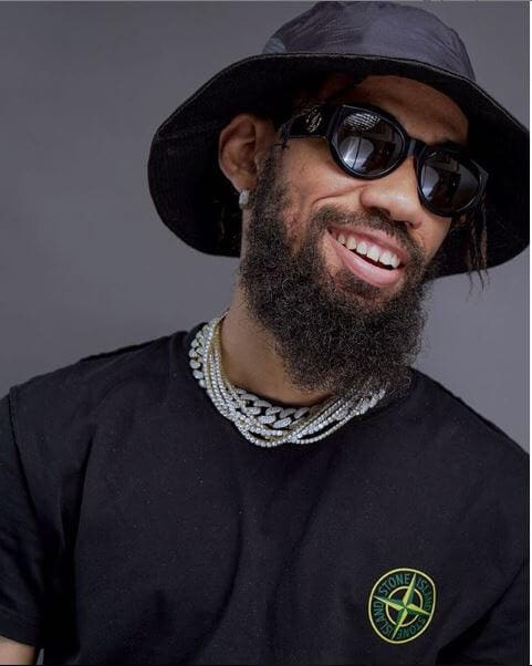 Phyno.jpg A man with a beard wearing a black hat, black sunglasses, and a black t-shirt with a circular emblem and multiple chain necklaces. He is smiling against a gray background, looking like he just dropped his new single.