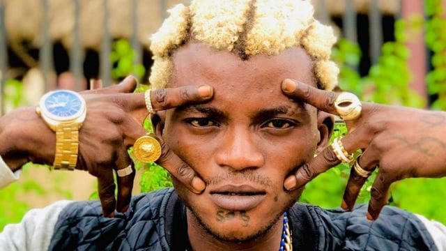 Portable.jpg A person with bleached hair and multiple gold rings poses with fingers framing their eyes. The individual also wears a gold wristwatch and a chain. The background shows greenery and a fence, capturing the essence of Naijaloaded where MUSIC LOVERS celebrate Moveable's Music Career.