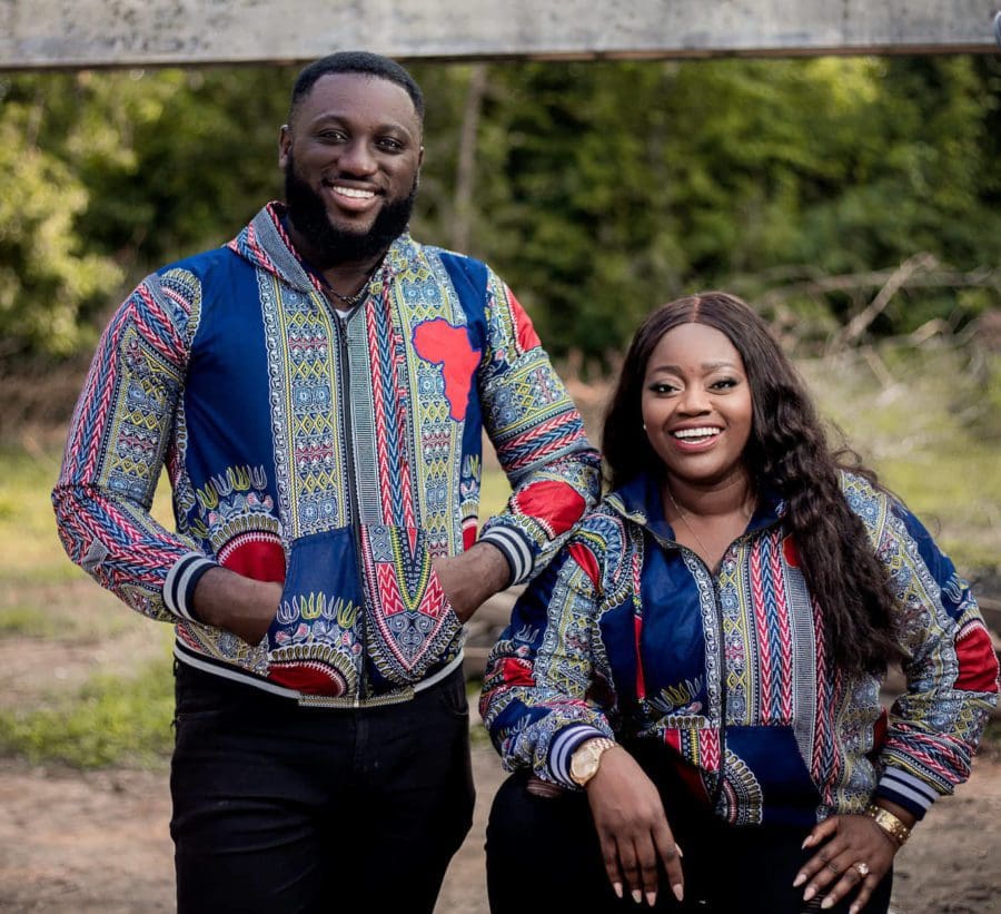 Sarah-sings-ft-MOG-Good-God-e1665741265482.jpg Two people smiling outdoors, both wearing multicolored, patterned jackets with similar designs. The person on the left stands with hands in pockets, while the person on the right sits, resting an arm on their knee. Their joyful expressions make it seem like they're enjoying some tunes from Afrobeat gospel artists like MOGmusic or Sarah Sings.