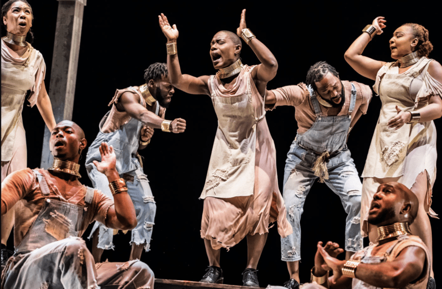 Screen-Shot-2022-10-26-at-9.48.57-PM-900x589.png Six performers in costume express emotions passionately on stage, wearing neutral-toned outfits with brass neck and arm pieces, against a dark background at the Cutler Majestic Theatre.