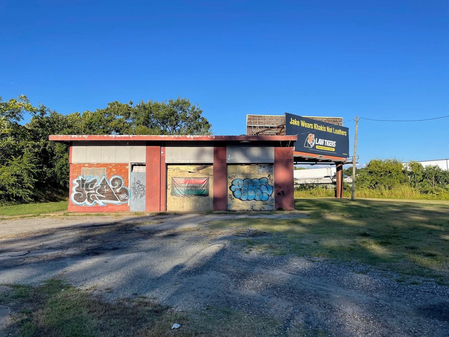 ZUOEYJLA3ID7ZCFWFKH72JSNW4.JPGw1440.jpeg An abandoned building with graffiti on the doors and walls stands in Richmond, surrounded by overgrown vegetation. A large billboard on top features an advertisement for land leases, reminiscent of a time when ancestors walked past what once might have been an African burying floor.