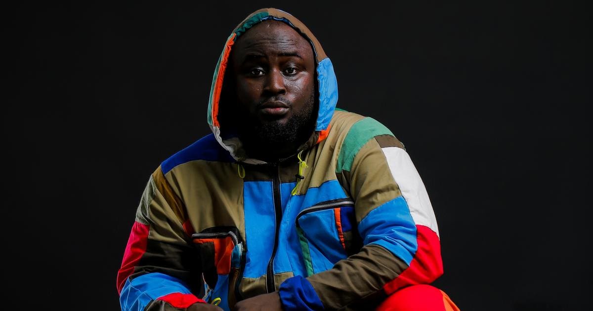 duXktkpTURBXy9jZmQzNTQwMzc4NzExZTM2NDg1MzBkZGViN2ExMjdmOS5qcGeSlQMAzQY_zQ8AzQhukwXNBLDNAnY.jpeg A man wearing a brightly colored patchwork hoodie and red pants, channeling That Unserious Focus Boi energy, sits against a black background.