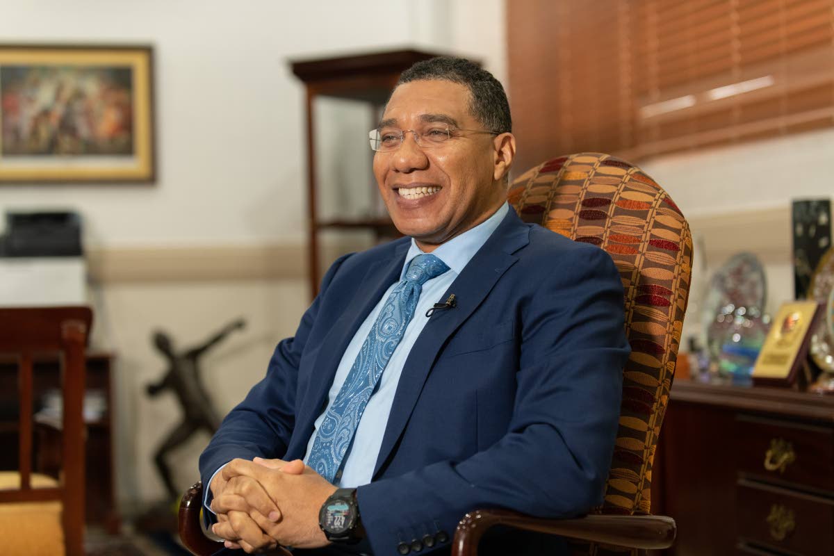 holness.jpg A man in a blue suit and tie sits in an office chair, smiling. The background includes various objects, such as framed artwork and awards, giving the room an air of achievement reminiscent of PM Holness's accomplishments.