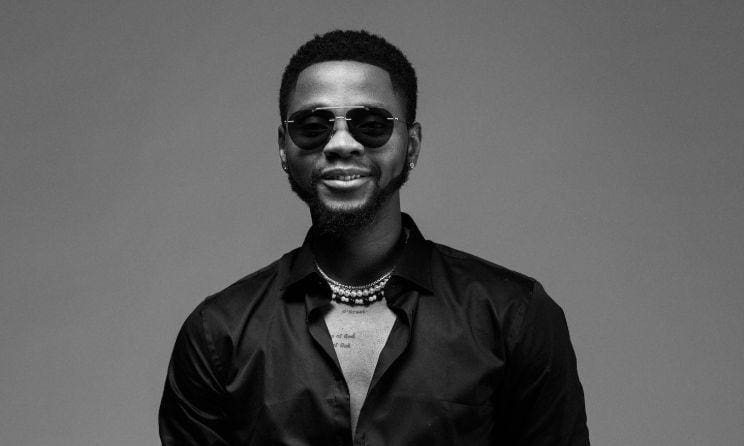kizzdaniel1.jpg A man wearing dark sunglasses, a Kizz Daniel necklace, and a black shirt smiles while looking at the camera in a black-and-white photo.