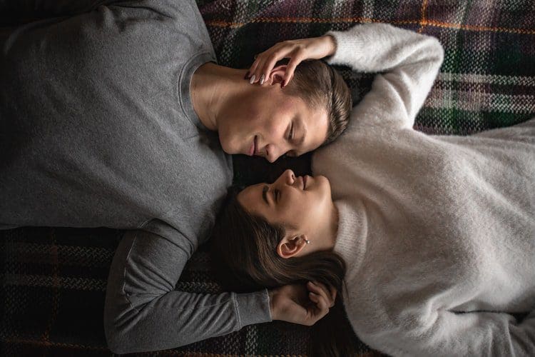 lovers.jpg A man and a woman lie on a blanket with their heads close together, each with one arm resting above their heads. They are both facing each other with their eyes closed, wearing gray and white sweaters respectively—resembling a serene moment after a lengthy time in bed.