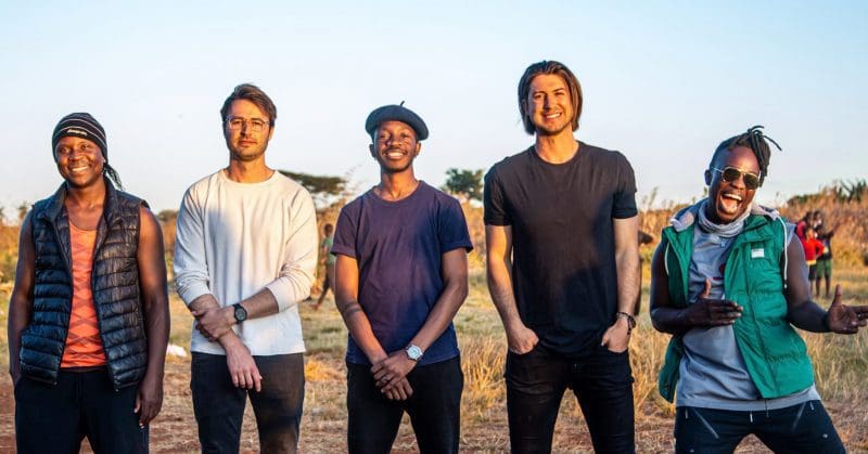 press-shot-scaled-e1665746776916.jpeg A group of five men standing outdoors, smiling. Three are wearing casual shirts, one is in a vest, and another in a green jacket with a headband. They embody the spirit of Africa And Kingdmusic, set against a natural backdrop under a clear sky.