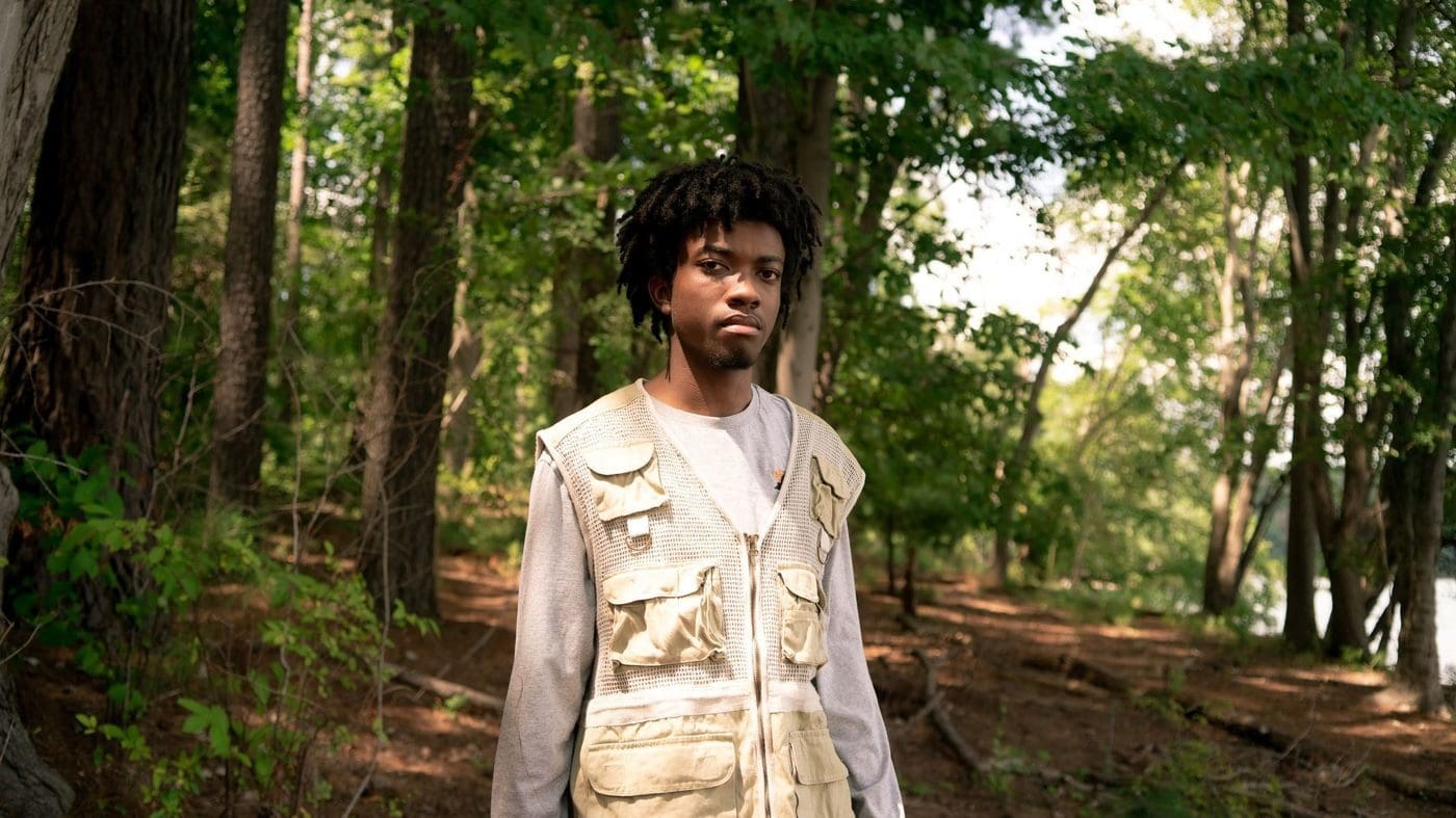 redveil_ethankwon_wide-289c7fce6c989a19949560f74d6bbafe5f7115b9-s1400-c100.jpg A person wearing a beige vest stands in a forested area with tall trees and sunlight filtering through the leaves, looking like they just stepped out of an Armani Caesar catalog.