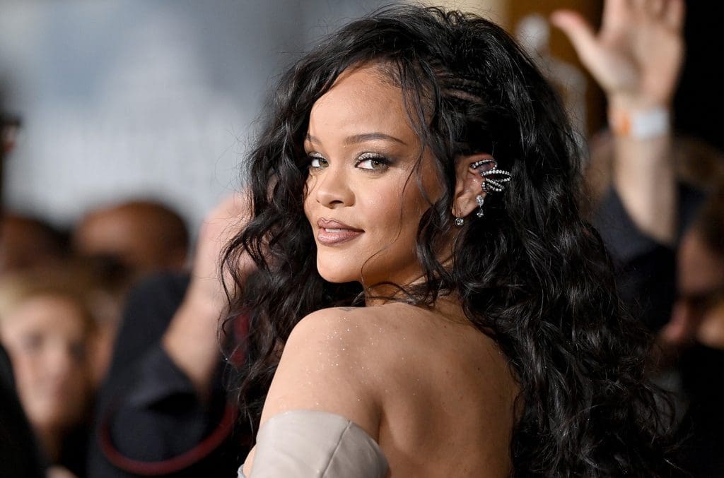 rihanna-wakanda-forever-2-rc-billboard-1548.jpg A woman with long black hair wearing an elaborate ear cuff and a shoulder-baring outfit looks over her shoulder, reminiscent of an iconic Rihanna billboard, surrounded by a crowd.