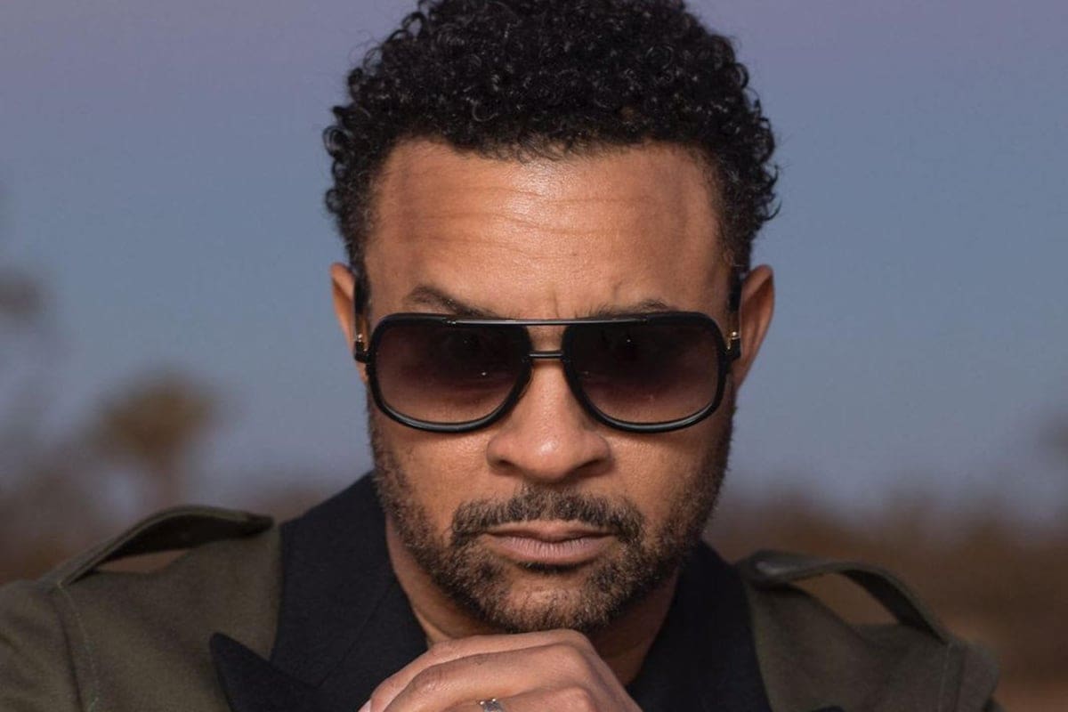 shaggy.jpg A man with curly hair and a beard is wearing dark sunglasses and a green jacket, looking directly at the camera at the Island Music Convention. The background is out of focus and appears to be outdoors.