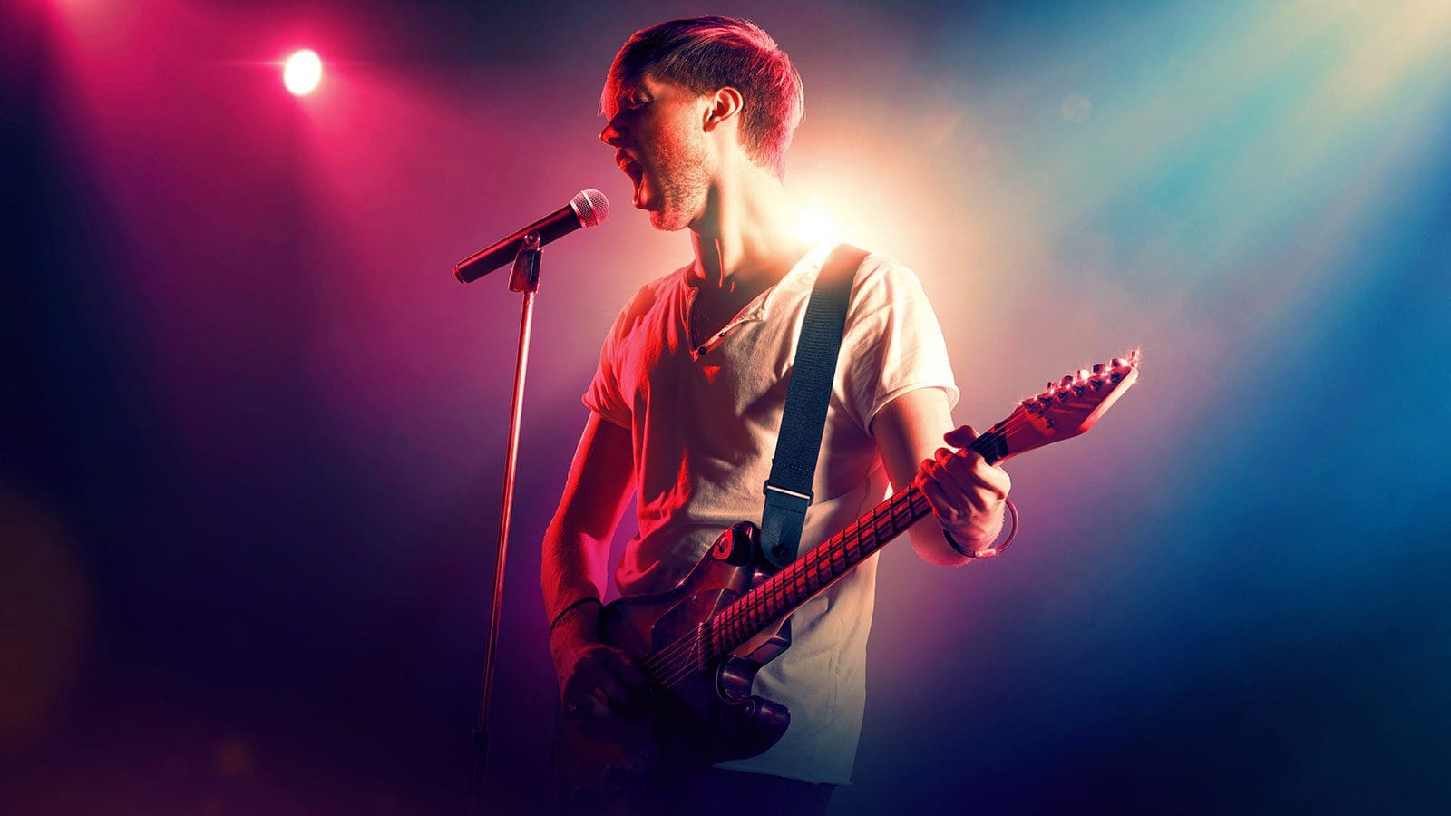 singer-guitar.jpg A musician in a white shirt plays the electric guitar and sings into a microphone on stage, illuminated by colorful stage lights, reminiscent of Louis Tomlinson's "Walls" video.