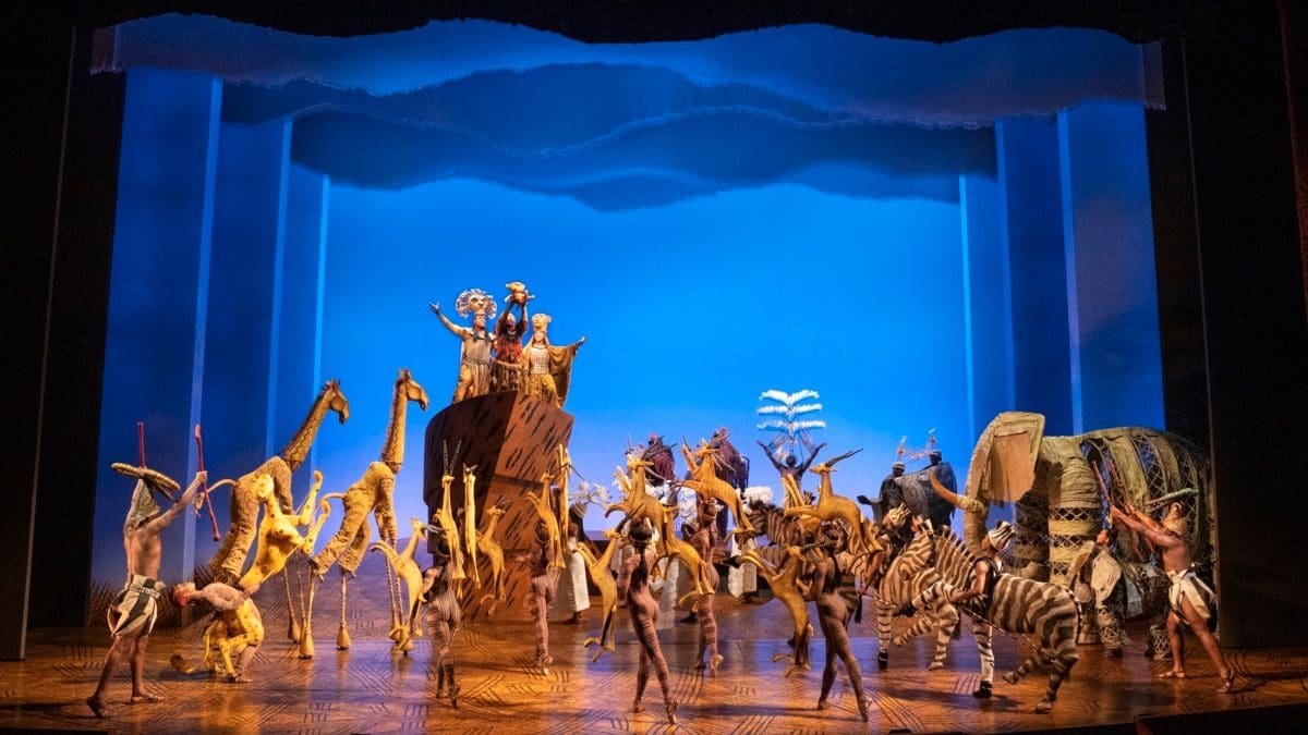09850000-0a00-0242-5a38-08dac978c87b_cx0_cy3_cw0_w1200_r1.jpg The theatrical stage is filled with performers in elaborate animal costumes during a musical production, reminiscent of Broadway's Lion King, set against a bright blue backdrop with cloud-like scenery.