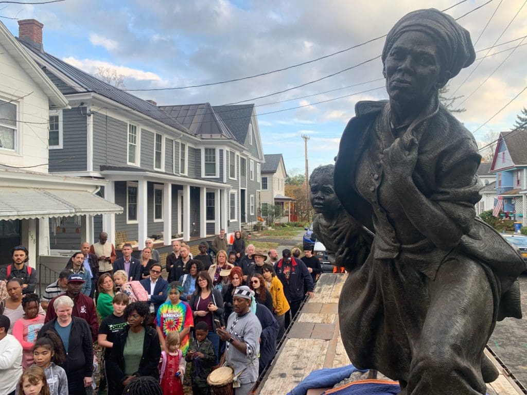 A crowd gathers around a large bronze statue of Harriet Tubman, dressed in period clothing, holding a child. The powerful Freeman figure stands proudly on a wooden platform in the heart of Kingston's residential neighborhood.