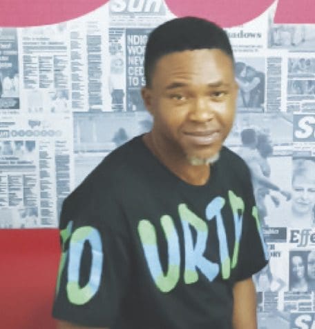 9-17.jpg A man with short hair and a goatee, wearing a black shirt with colored text that hints at being a fan of an American rap star, is seated in front of a wall covered with newspaper clippings.