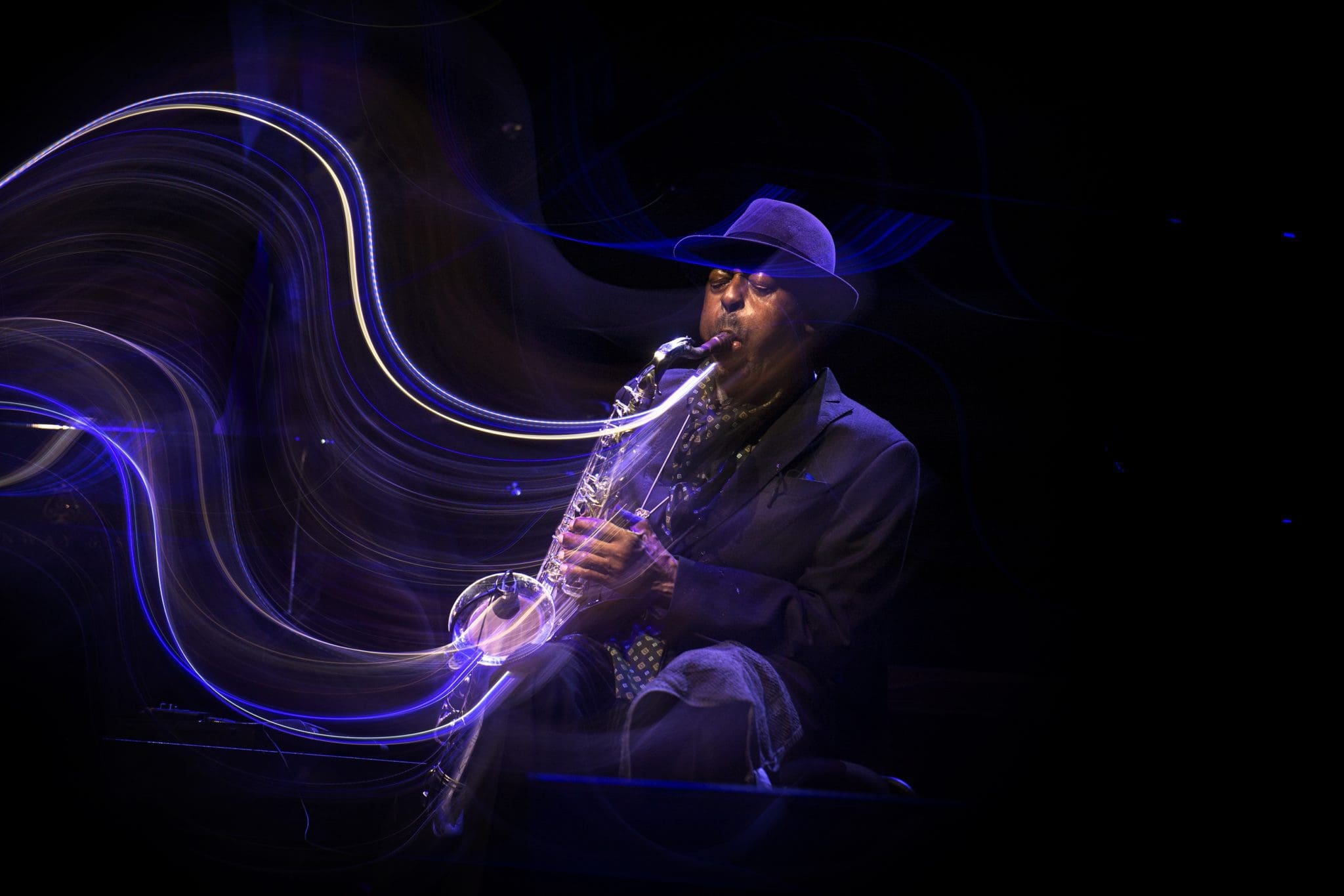 Archie-Shepp-Barbican-by-Tatiana-Gorilovsky4_lines_2021-11-25-160156_bokl-scaled.jpg A person in a dark suit and hat mesmerizes the audience at the London Jazz Competition with their saxophone performance, accompanied by swirling light effects.