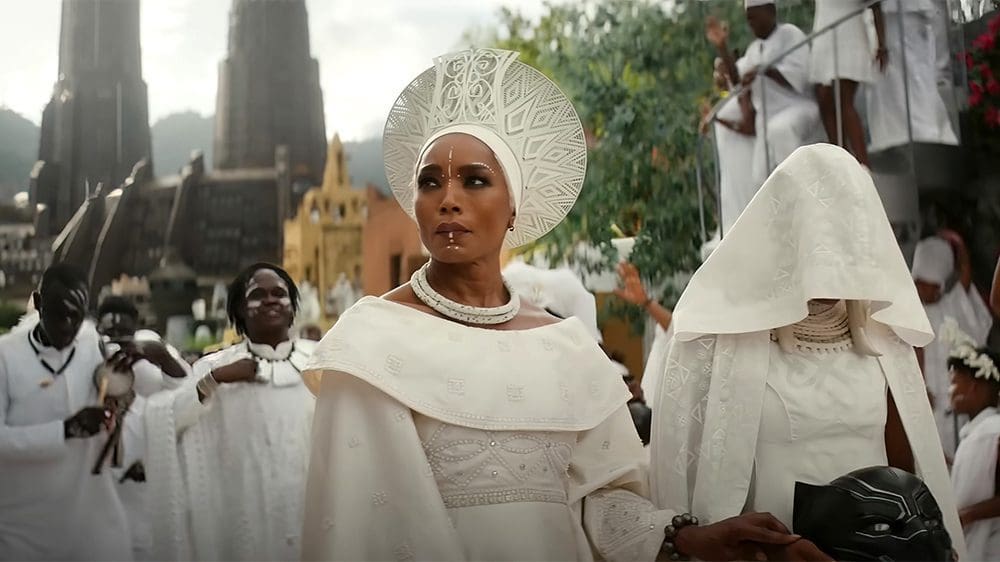 Black-Panther-3.jpg A person dressed in ornate white attire and headdress stands in a procession with others clad in similar white garments, set against an outdoor background with buildings and greenery, reminiscent of the cultural richness seen in Wakanda.