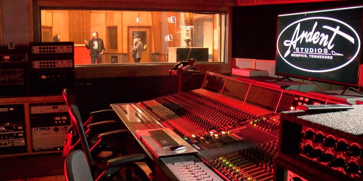 DCCM54NKJVE3ZJ5HI4RXNUOBJM.png View of a recording studio control room with a mixing console in the foreground, a large window showing people in the recording booth, and a monitor displaying "Ardent Studios" on the right. The music-infused atmosphere captures creativity at its peak.