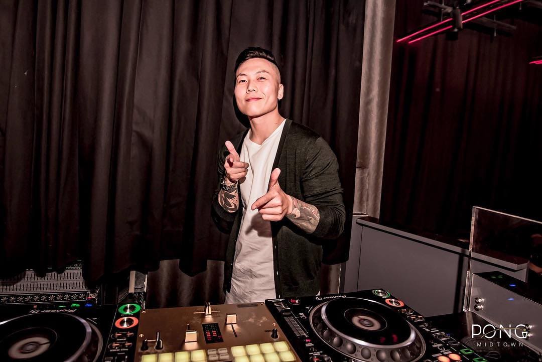 DJ-Sidtrus.jpeg A DJ standing behind his equipment, pointing towards the camera with both hands. He is in a dimly lit room with dark curtains in the background, announcing DJ Sidtrus's new album collaboration with Yung Bos.