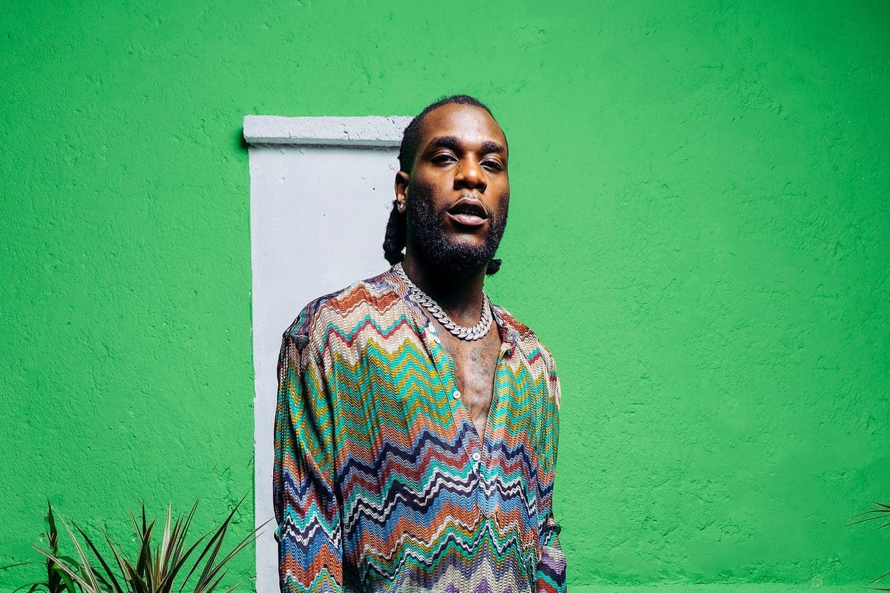 burna.jpg A man stands in front of a vibrant green painted wall, wearing a colorful zigzag patterned shirt and a silver chain necklace, reminiscent of the energetic vibe you’d find at a Burna Boy live performance in Jamaica.