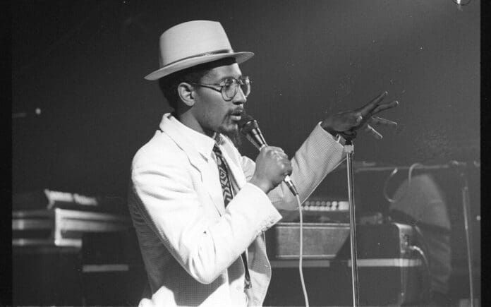 Linton-Kwesi-Johnson-e1667475596925.jpeg A man wearing a hat and glasses sings into a microphone on stage, gesturing with his hand. He is dressed in a white suit, reminiscent of the iconic style of Linton Kwesi Johnson during one of his memorable five dubs performances.