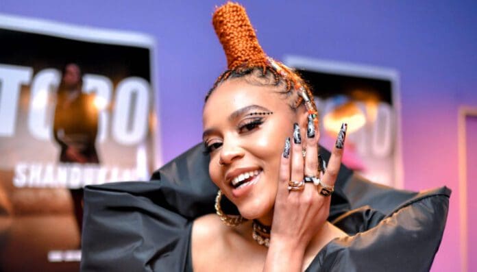 image_6483441-5-scaled-e1669639869199.jpg Person smiling and touching their face with long, decorated fingernails. They have braided hair styled in a high Mohawk and are wearing a black outfit with shoulder embellishments. Posters of Sho Madjozi are in the background, reflecting South Africa's vibrant culture featured in The Africa Report.