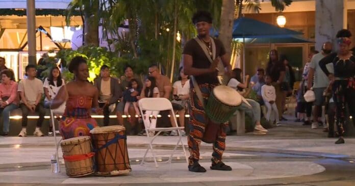 kwanzaa-celebration-at-the-square-in-west-palm-beach.JPG Two drummers perform outdoors, one sitting and one standing, as a crowd watches from behind. Surrounded by trees and evening lights, the rhythmic music sets a perfect backdrop for the Kwanzaa celebration.