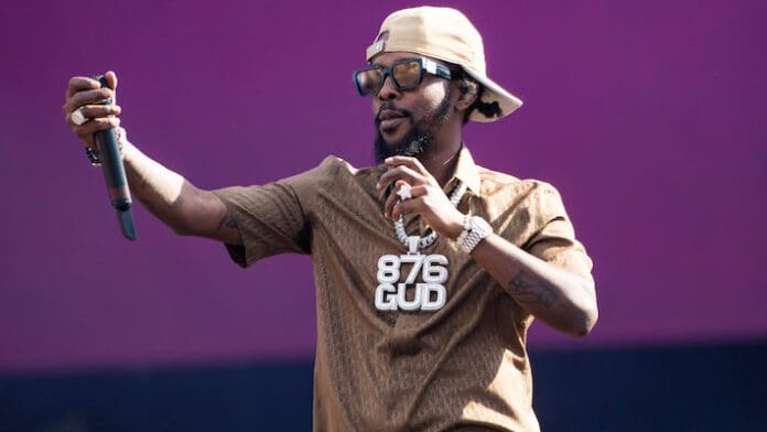 2-popcaan.jpg A man wearing a tan shirt and cap, large sunglasses, and a necklace with "876 GUD" pendant holds a microphone with an outstretched hand against a purple background. Could this be Popcaan's new single featuring Drake?