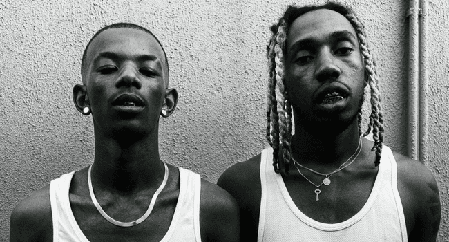 Two people stand side by side against a textured wall, both wearing white tank tops and necklaces. One with braided hair and the other with short hair, they exude an aura reminiscent of the vibrant energy of Maglera Doe Boy’s next music concept.