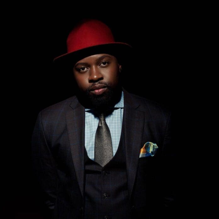 Mr-JazziQ-–-Stoff-ft.-Busta-929-1.jpg A man wearing a red hat, blue checkered shirt, dark suit, and tie stands against a dark background as if ready to perform alongside Mr JazziQ.