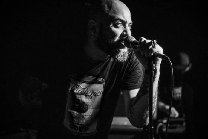 Scuru-Fitchadu-1010x673.jpeg A man with a beard and tattoos sings into a microphone on stage, wearing a T-shirt with a can graphic. The image is in black and white, capturing the essence of Scúru Fitchádu's energetic fusion of funaná and Pan African Music.