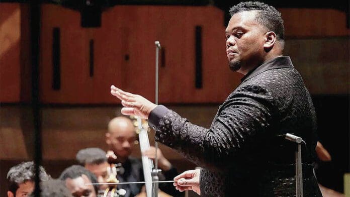 web1_20230129-d1-Anthony-Parthner-2-1-.jpg A conductor in a dark patterned suit is leading an orchestra through a classical music performance. The conductor holds a baton and has his left hand raised, guiding the musicians with various instruments in the background. This captivating performance is taking place in February in Honolulu.