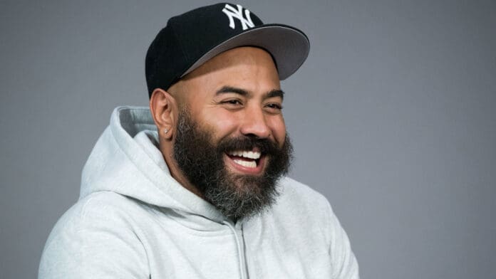 Ebro-Darden.jpeg A smiling bearded man sporting a black baseball cap and a light gray hoodie, looking as if he just finished jamming to some energetic Afrobeats, stands against a plain gray background.