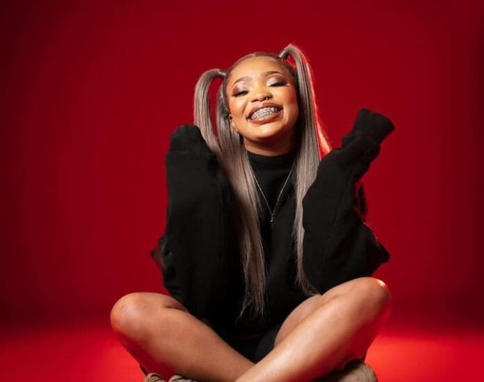 Babalwa M sitting on a red background with her hair in a ponytail.
