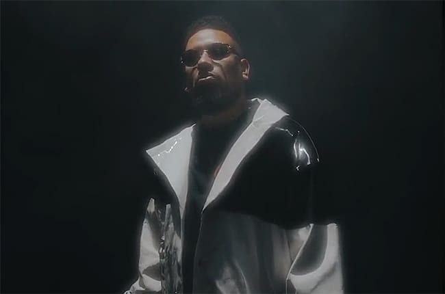 A man in sunglasses stands in the dark wearing a black jacket. A man in sunglasses stands in the dark wearing a black jacket.
