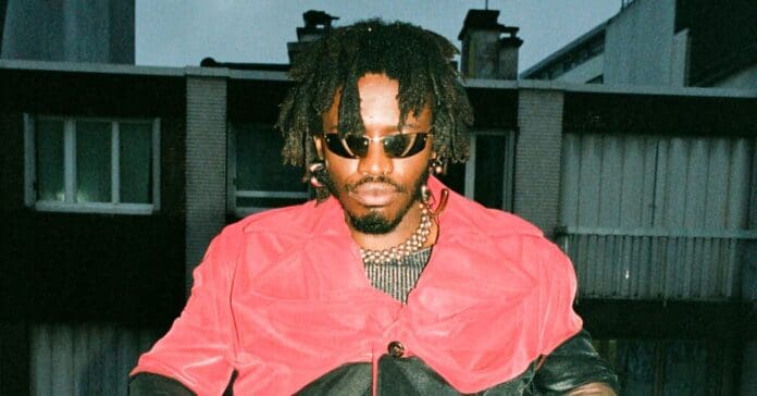 Desire Marea, a South African artist known for their genre-melting music, stands out with their iconic dreadlocks and stylish red jacket, completing the look with fashionable sunglasses. Desire Marea, a South African artist known for their genre-melting music, stands out with their iconic dreadlocks and stylish red jacket, completing the look with fashionable sunglasses.
