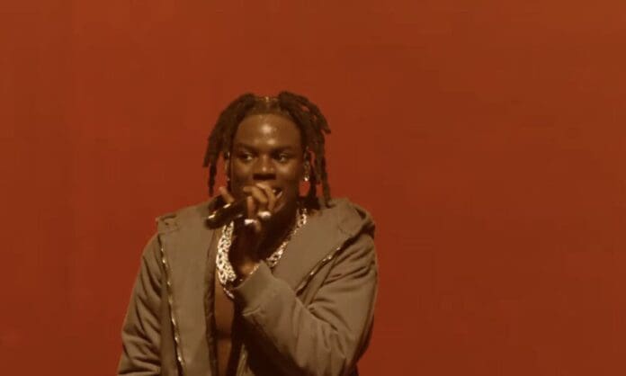 Rema, a man with dreadlocks, is holding a microphone on stage at the O2 Arena concert. Rema, a man with dreadlocks, is holding a microphone on stage at the O2 Arena concert.