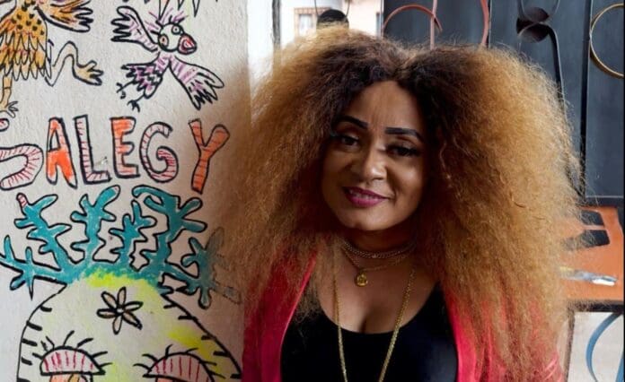 0066172593ca1850b212ebdb05734c1d04e54176arc614x376w1200.jpg A person with voluminous curly hair stands next to a wall adorned with colorful graffiti, featuring the word "Salegy"—a nod to vibrant Malagasy music from Madagascar.