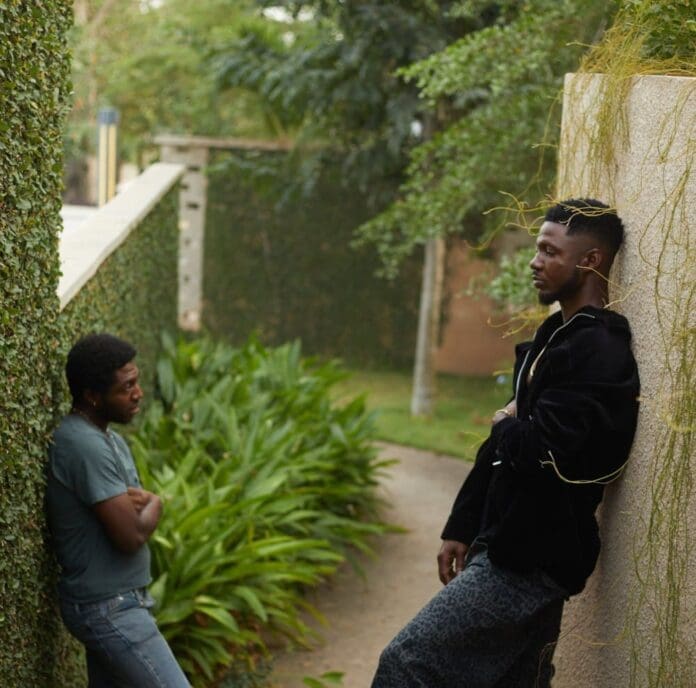 Two people stand in an outdoor garden corridor, one leaning against a wall, the other resting against a hedge. The rhythm of Afrobeats plays softly in the background, making the moment feel even more precious.