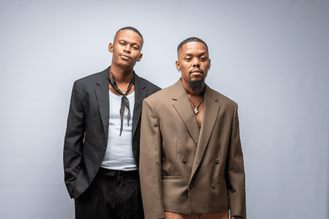 Two individuals stand side by side in stylish ensembles, as if ready for a feature on TimesLIVE. The one on the left sports a black blazer, white shirt, and scarf, while their companion dons a brown blazer paired with a statement necklace.