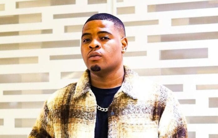 IMG_20230712_122040-e1741597327169.jpg A person with short hair dons a patterned jacket and a chain necklace, standing strikingly before a geometric backdrop reminiscent of Bona Magazine's bold style, evoking vibes of amapiano energy.