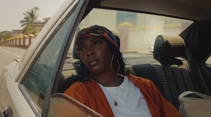 Tiwa-savage-water-and-garri.webp.webp In the car's back seat, a woman in an orange jacket and headscarf sits, gazing out the open window on a sunny day, feeling the rhythm of Tiwa Savage's music as it weaves through her thoughts.